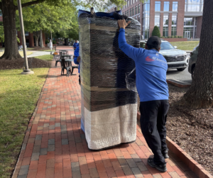Two movers wrap a large rectangular item as they move it along a brick-paved path.